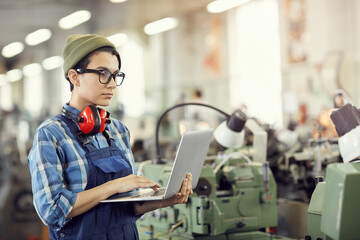 Wall Mural - Serious focused young female industrial engineer in beanie hat and eyewear standing in factory shop full of lathes and using laptop