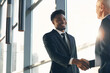 © Mediaphotos - Cheerful handsome young Black businessman in suit standing at panoramic window and making handshake with business partner in lobby of company