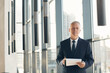 © Mediaphotos - Portrait of serious senior business analyst with gray hair standing at panoramic window and holding clipboard with papers