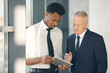© Mediaphotos - Serious mature businessman in formal suit pointing at digital tablet while explaining data to young Afro-American colleague