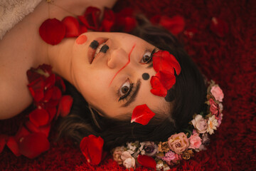 Close portrait of woman lying on a red furry textured rug with her face painted with an alternative makeup and with rose petals covering her face and a colorful flower crown in her hair