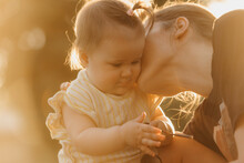 A Female Toddler In Yellow Clothes Is Playing With Sunglasses Near Her Mom In The Sun Rays. Mother Is Kissing Her Young Daughter In The Park At Sunset.