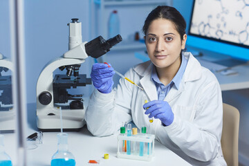 Portrait of content young Indian female pharmacist in lab coat and surgical gloves sitting at desk and mixing reagents in test tube using pipette