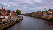 York, UK. People Walk By The Embankment Area Of The Ouse River In York, UK. Time-lapse Of Heavy Sunset Clouds And Touristic Boats. Cloudy Evening, Zoom In