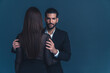 Studio shot on a dark blue background. Brunette long-haired woman standing back to camera, latin unshaved elegant man looking at camera, holding the woman by her shoulders. High quality photo