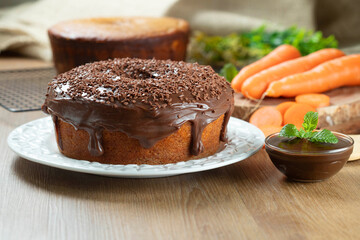 Wall Mural - Brazilian carrot cake with chocolate frosting on wooden table with carrots in the background