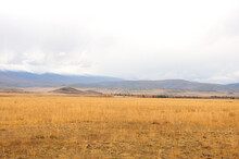 Panoramic Shot Of A Dried Up Hilly Steppe In Early Autumn At The Foot Of A Mountain Range Under An Overcast Sky.