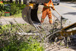 Closeup view on hydraulic arm of an excavator at work clearing tree branches and debris from the highway after a storm, man with rake in background.