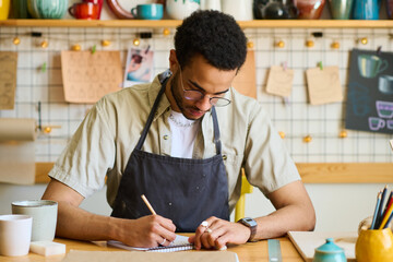 Aufkleber - Young man in casual shirt and black apron drawing sketch of clay item in notepad while working over new collection of handmade products