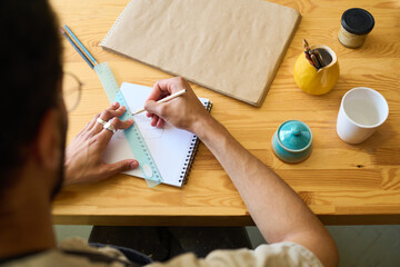 Aufkleber - Above view of hands of young man with pencil and ruler drawing sketch of new creative earthenware item on page of notepad by table