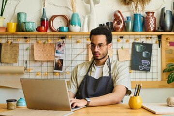 Aufkleber - Young serious black man in workwear and eyeglasses sitting by workplace in front of laptop and looking for new online orders of clients