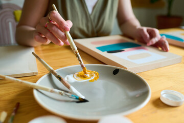 Aufkleber - Hand of young creative female with paintbrush mixing white and yellow colors on plate while sititng by table and painting on paper