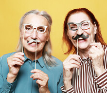 Funny Elderly Female Friends With Fake Mustache And Glasses, Laughs And Prepares For Party Over Yellow Background