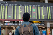 Back of young male traveler with backpack wearing a cap and looking at departure board in airport terminal. Male tourist checking flight time table on board - with copy space