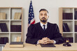 © Studio Romantic - Serious incorruptible young judge dressed in a robe uniform sitting at his table with books and gavel in the courtroom during a court hearing in an American courthouse. US law and justice concept