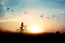 Lonely Woman Standing With Bicycle On Road Of Paddy Field Among Flying Birds And Sunset Background