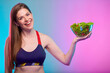 Smiling sporty woman in fitness sportswear holding glass bowl with green salad  Female fitness portrait isolated on neon multicolor background. Healthy food.