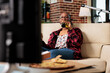 © DC Studio - Cheerful adult drinking alcoholic beer from bottle on couch, watching movie or film on television channel. Eating fast food meal from delivery package and enjoying beverage in living room.