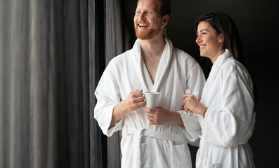 Poster - Happy young couple in white bathrobes drinking coffee together. Hotel, travel, relationships concept