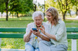 © Halfpoint - Adult granddaguhter helping her grandmother to use cellphone when sitting on bench in park in summer.