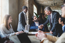 Two Businessmen Make A Deal By Shaking Hands Sitting At A Table Together With Other Multiracial Employees - Business Lifestyle And Agreements Concept