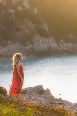 Blond woman wearing a loose red dress standing in profile at the edge of the hair blowing in the wind looks at the sea.  Sunset light with flair. Image with soft color toning.