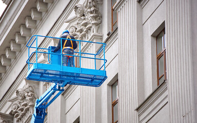 Construction worker in lift bucket of crane restore and repair historic facade of building. Man in cradle, restoring plaster decoration on facade. Workers painting building, renovating and repair work