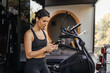 © Look! - Pretty young caucasian girl is typing message on phone standing in gym. Brunette wears black sports top and leggings. Social media addiction concept