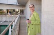 © KAMPUS - Smiling grey-haired businesswoman in mall. Woman in suit with ponytail drinking coffee. Portrait, business concept