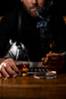 © fesenko - view on male hand holding glass with cognac and smoking cuban cigar over ashtray