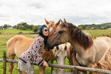 A Beautiful Girl In Glasses And With A Bandana On Her Head Strokes The Horses In The Meadow. Blurred Background.