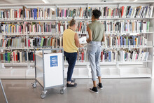 Female Librarian Helping College Student With Books In Library