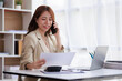© David - Portrait of a young Asian woman sitting at an office desk with a laptop computer and files and talking on a mobile phone. Communication concept