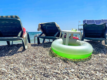 Vacation By The Sea On The Beach. On The Pebbly Ground There Are Sun Loungers For Sunbathing Tourists. Nearby Are Swimming Accessories, A Rubber Life Buoy For A Child
