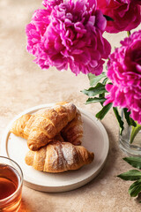 Plate of fresh croissants, tea and peonies flowers for breakfast on brown background with text space