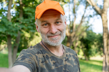 Self portrait of mature man wearing orange cap  standing in park outdoors. Senior caucasian man with grey beard taking selfie