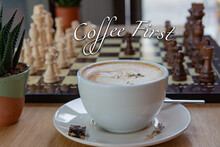 A Cup Of Frothy Coffee And Black And White Chess Pieces Lined Up On A Board In The Background. Inscription: COFFEE FIRST On The Top Of Image.