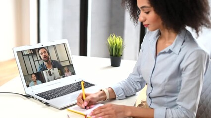 Wall Mural - Intelligent mixed-race businesswoman sitting at the desk and talking via video call with a group of diverse colleagues via laptop, having a virtual online meeting, taking a notes, writing in notepad