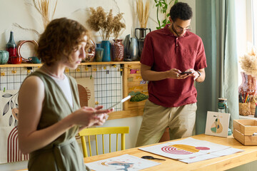Aufkleber - Young multi-ethnic man with smartphone taking photo of drawing on paper while standing by table with his colleague using tablet in front