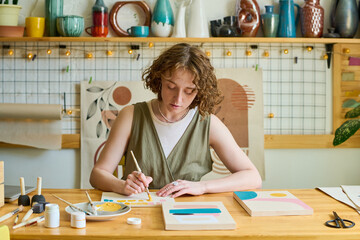Aufkleber - Young creative female in casualwear with paintbrush and gouache creating new painting while sitting by workplace in studio