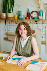 Aufkleber - Young successful woman with curly hair looking at camera while sitting by workplace with abstract paintings created by her for sale