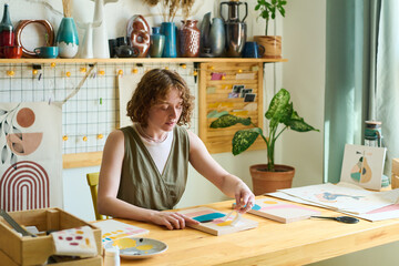Aufkleber - Young creative female with paper stencil creating abstract painting while sitting by table in studio with various supplies for artworks
