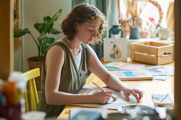 Aufkleber - Serious young woman with paintbrush sitting by workplace in studio and painting abstract stuff with watercolors or gouache on paper