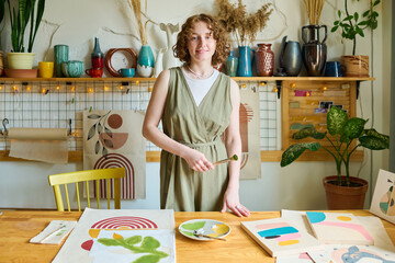 Aufkleber - Happy young creative woman in grey casual dress holding paintbrush in hand while standing by table with her drawings in workshop