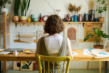 Aufkleber - Rear view of young creative woman sitting by table in workshop with handmade souvenirs on shelf along wall and creating new paintings