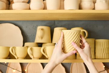 Aufkleber - Hands of young woman holding handmade clay mug while choosing one from assortment on shelves with other earthenware in shop