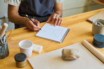Aufkleber - hands of young creative craftsman making sketch of cup on page of notepad while sitting by table with supplies for creating clay items