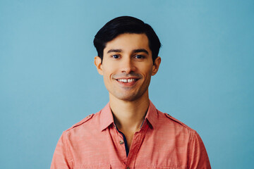 Headshot hispanic latino man black hair smiling handsome young adult wearing pink shirt over blue background looking at camera studio shot