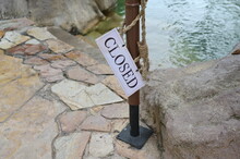 A White "Closed"  Sign Written In Black Ink Hangs On A Manila Rope Beside A Brown Steel Pole. The Closed Sign Has Been Take Off, Meaning Access To The Pool Is Possible.
