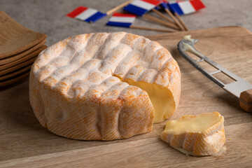 Wall Mural - Aged French soft cheese with orange washed rind from the Vosges mountain range on a cutting board close up with french flags in the background 